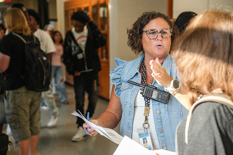 A woman with glasses holds papers and speaks to another person. Others in the background walk through a hallway.