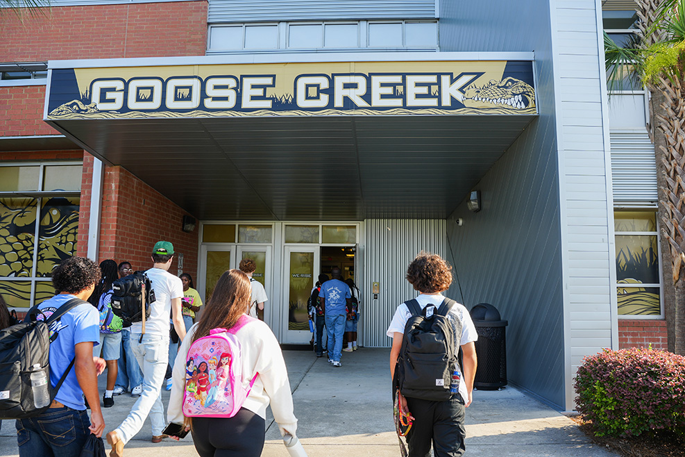 Students walk past an entrance sign reading "Goose Creek" with a building and palm tree in the background.
