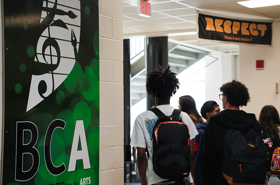 Several students walk in a corridor with a wall banner featuring "BCA" and a musical note.