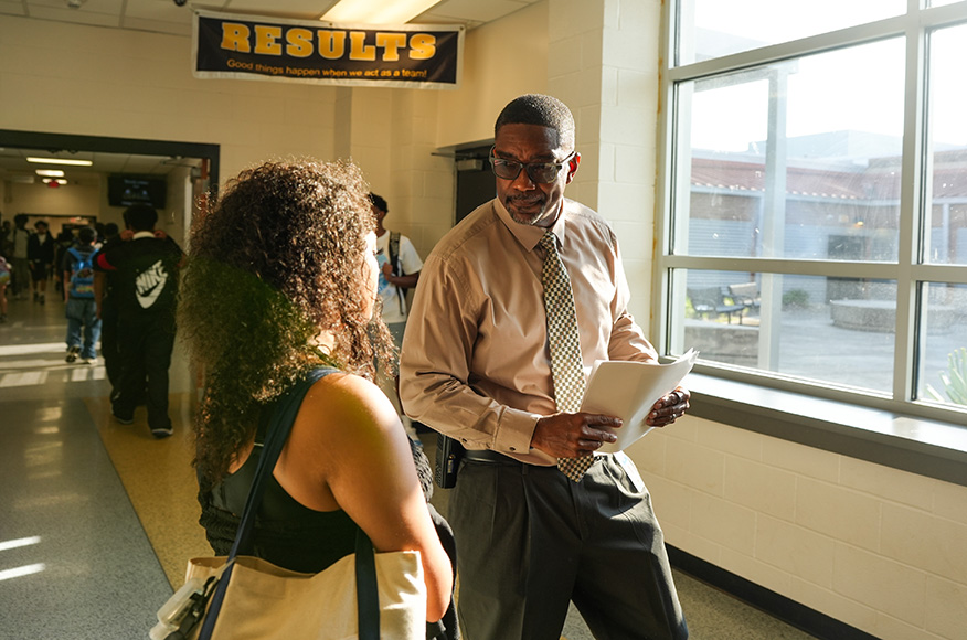 Two people conversing in a school hallway. A man with a tie holds papers, and a woman has a bag.