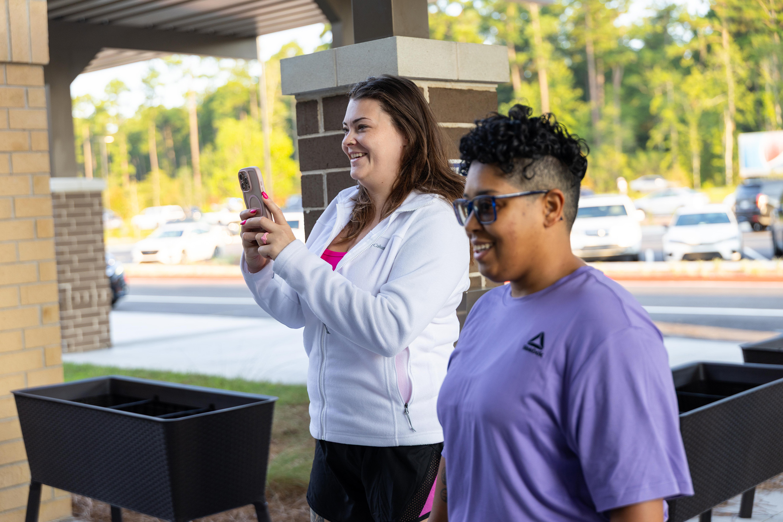 Two women stand outside; one holds a phone, and the other wears glasses. They are in front of a brick column.