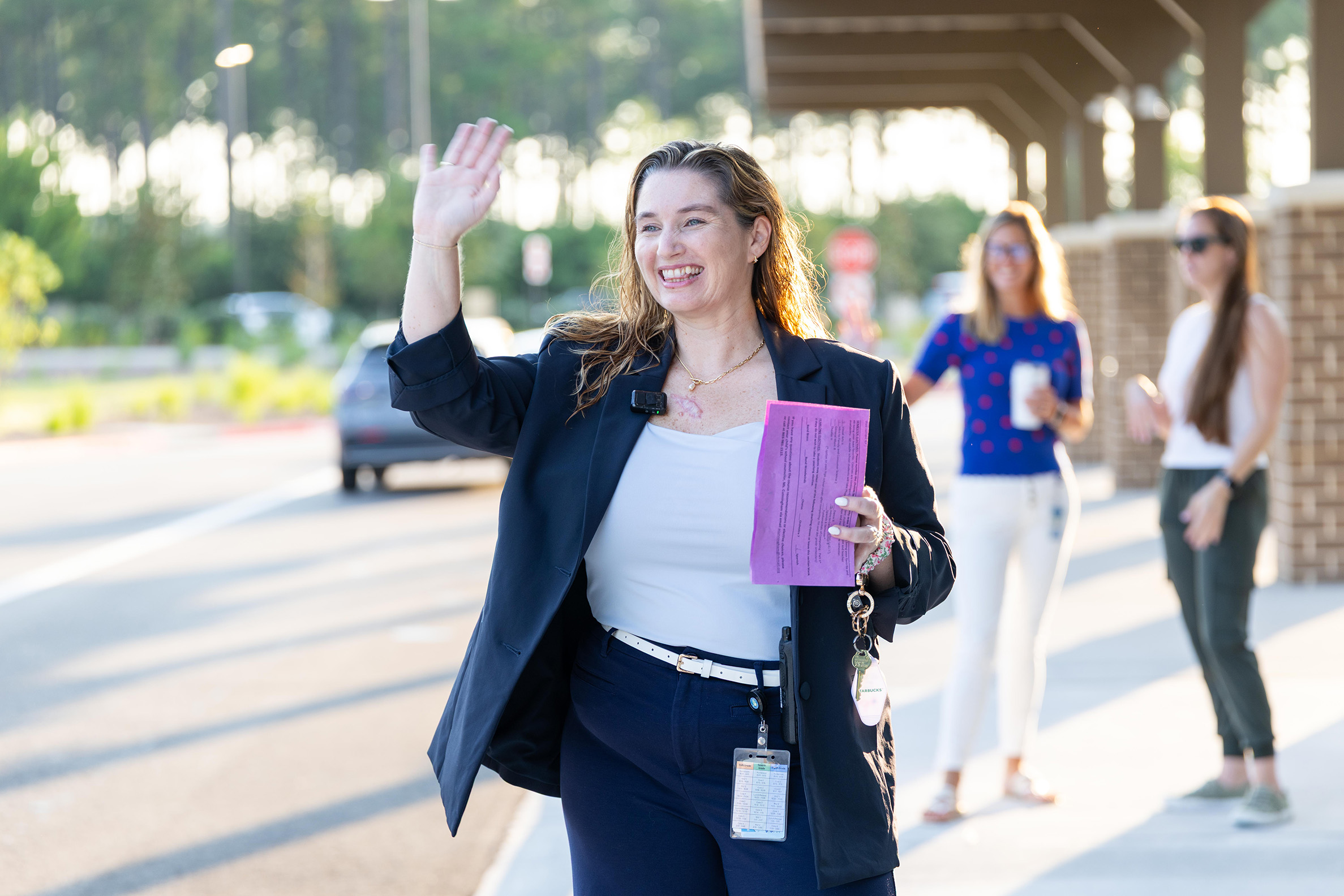 Woman in blazer waves while holding folder, another holds drink. Two women in background walk on sidewalk.