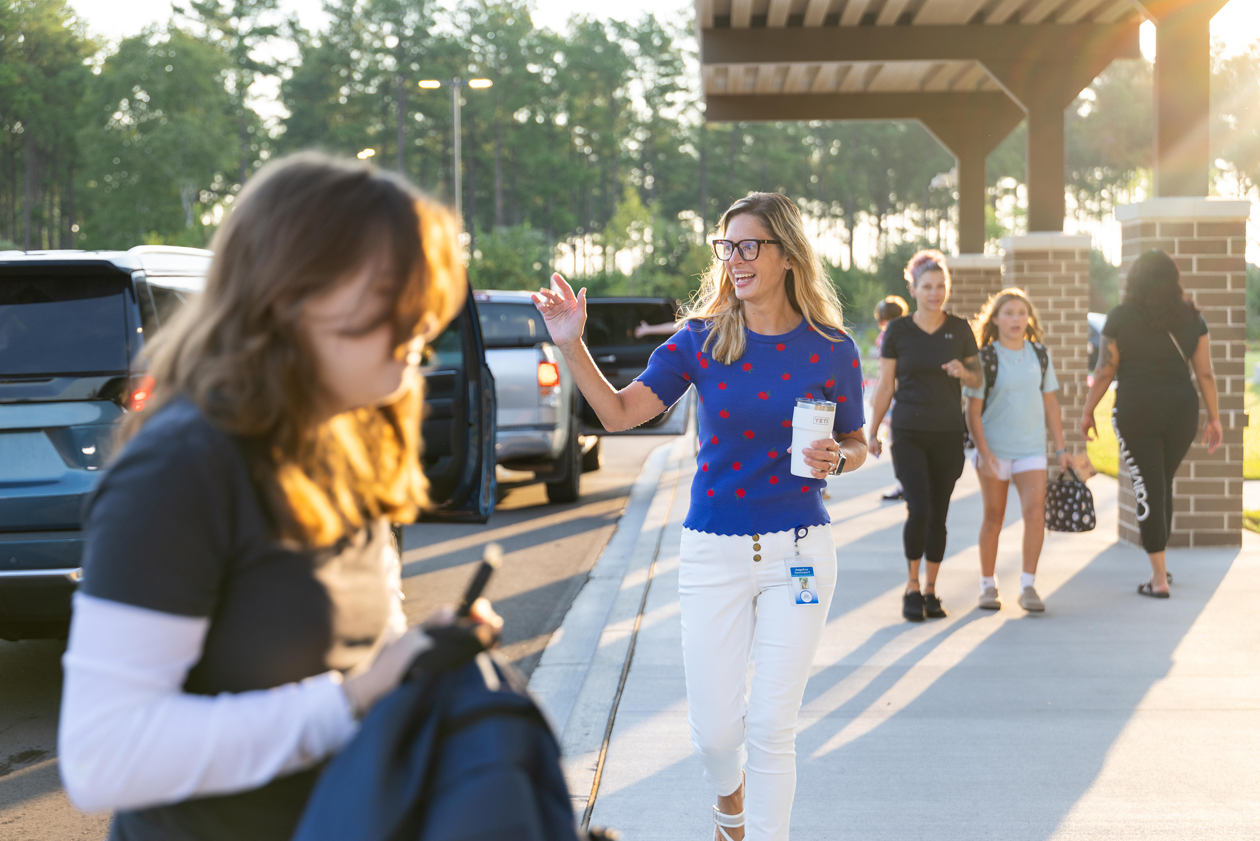 People walking on a sidewalk, one waving. Cars parked behind them. One person holds a cup. Trees and a building in the background.