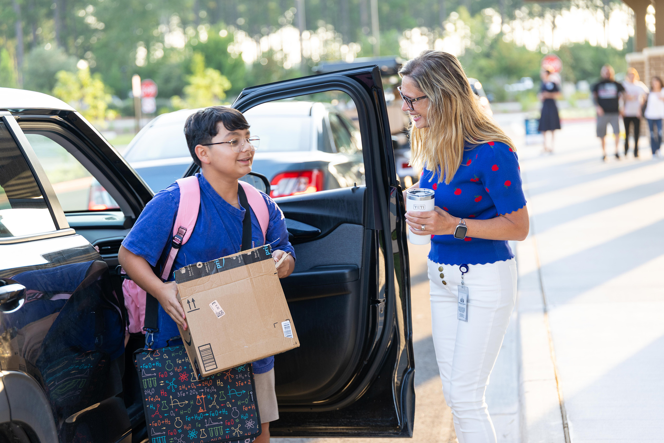 A person with glasses and a backpack helps a person with a box while another person holds a drink.