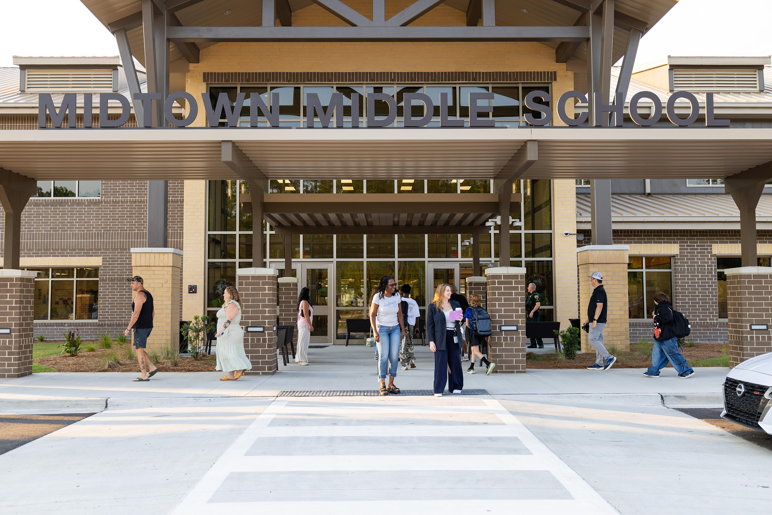 A school entrance with a large sign reading "MIDTOWN MIDDLE SCHOOL." Several people walk past it.