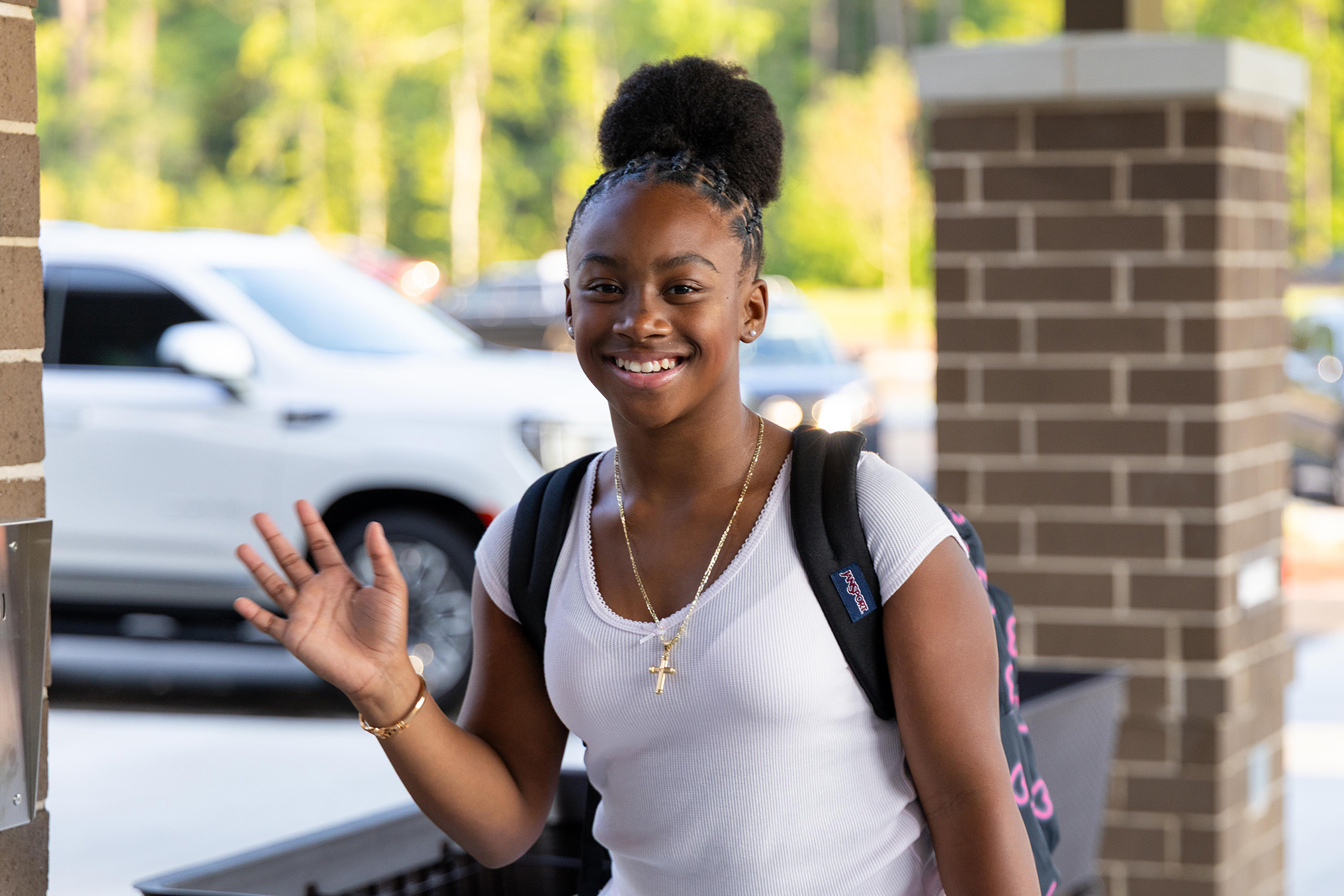 Person wearing white t-shirt and backpack waves to camera. Brick pillar behind them. Car parked in background.