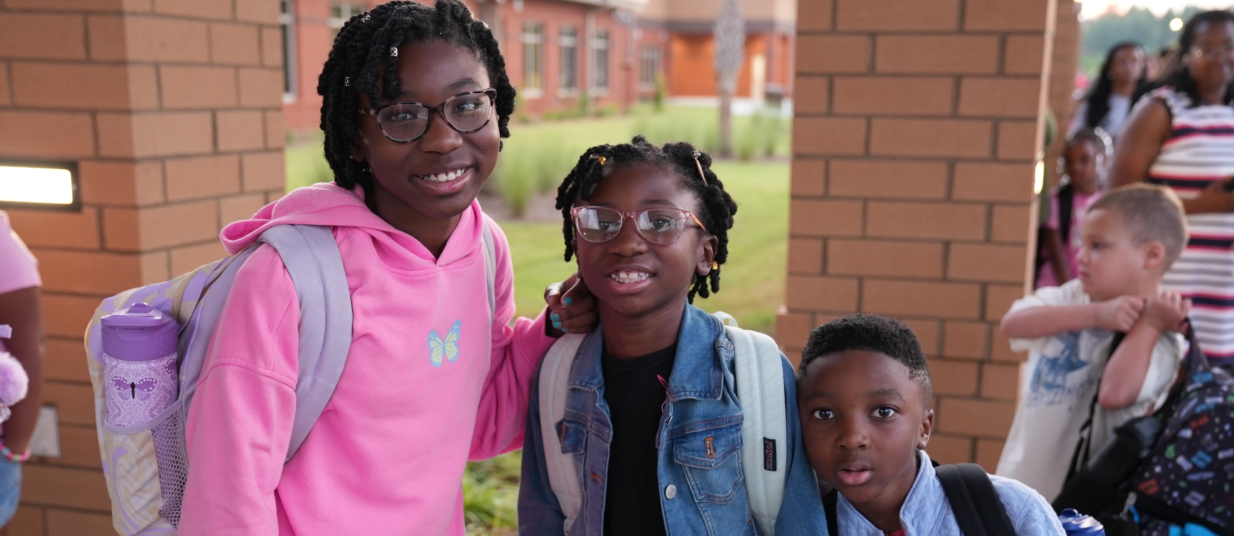 Three children pose for a photo; a girl in pink, a girl in denim, and a boy in blue.