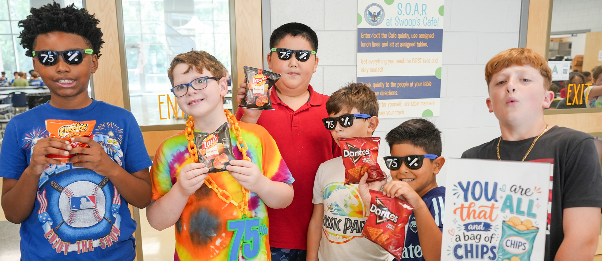 A group of children wearing sunglasses holds snacks, standing in a hallway. A sign behind them says, "YOU are that ALL of CHIPS."