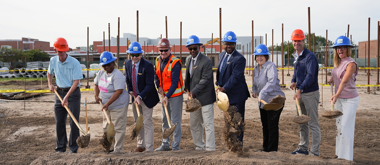 Nine people in helmets and hard hats hold shovels and stand around a dirt mound at a construction site.