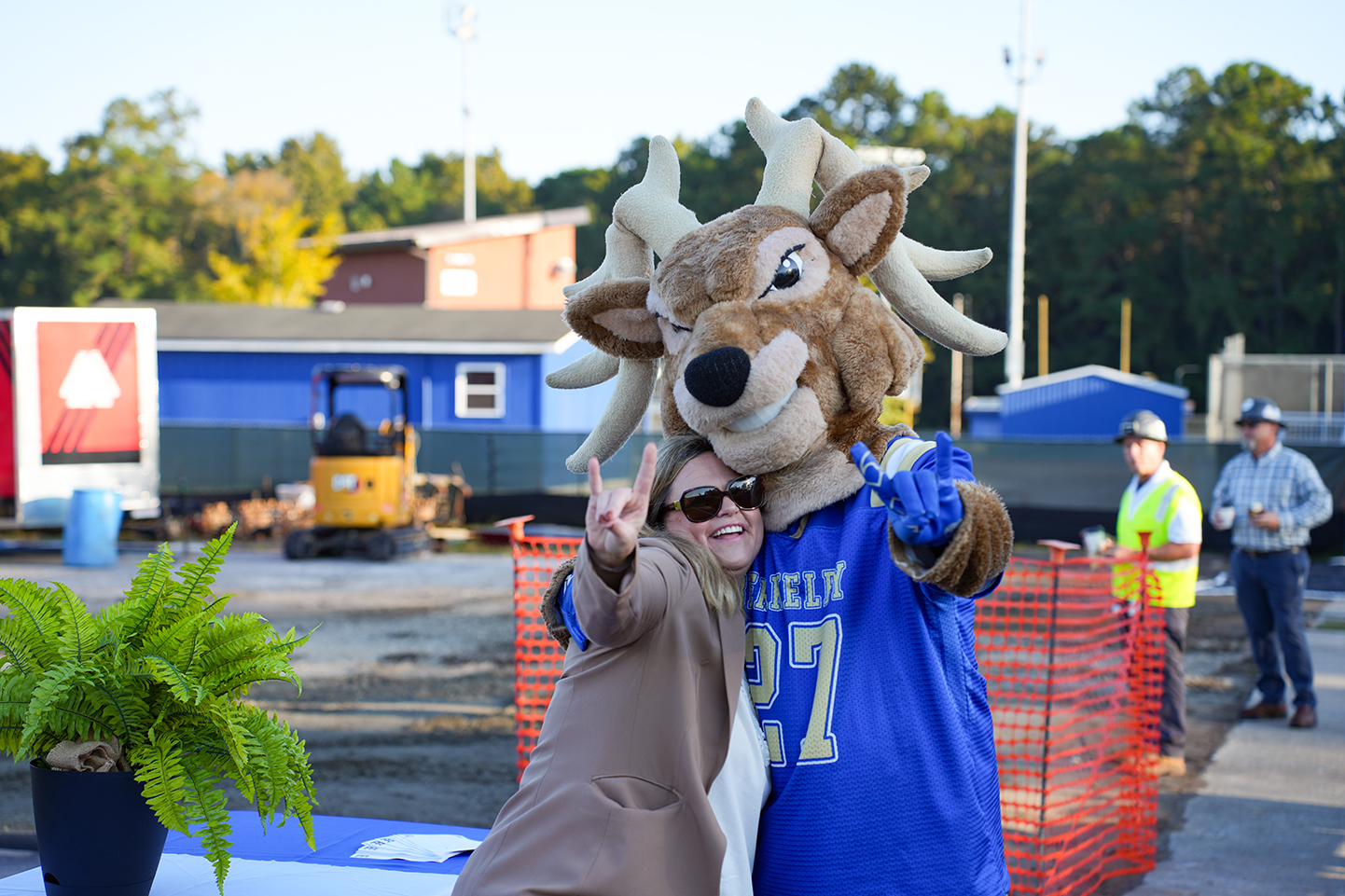 Person and mascot in blue jersey pose for photo with finger guns. Behind them are plants, barriers, and construction vehicles.