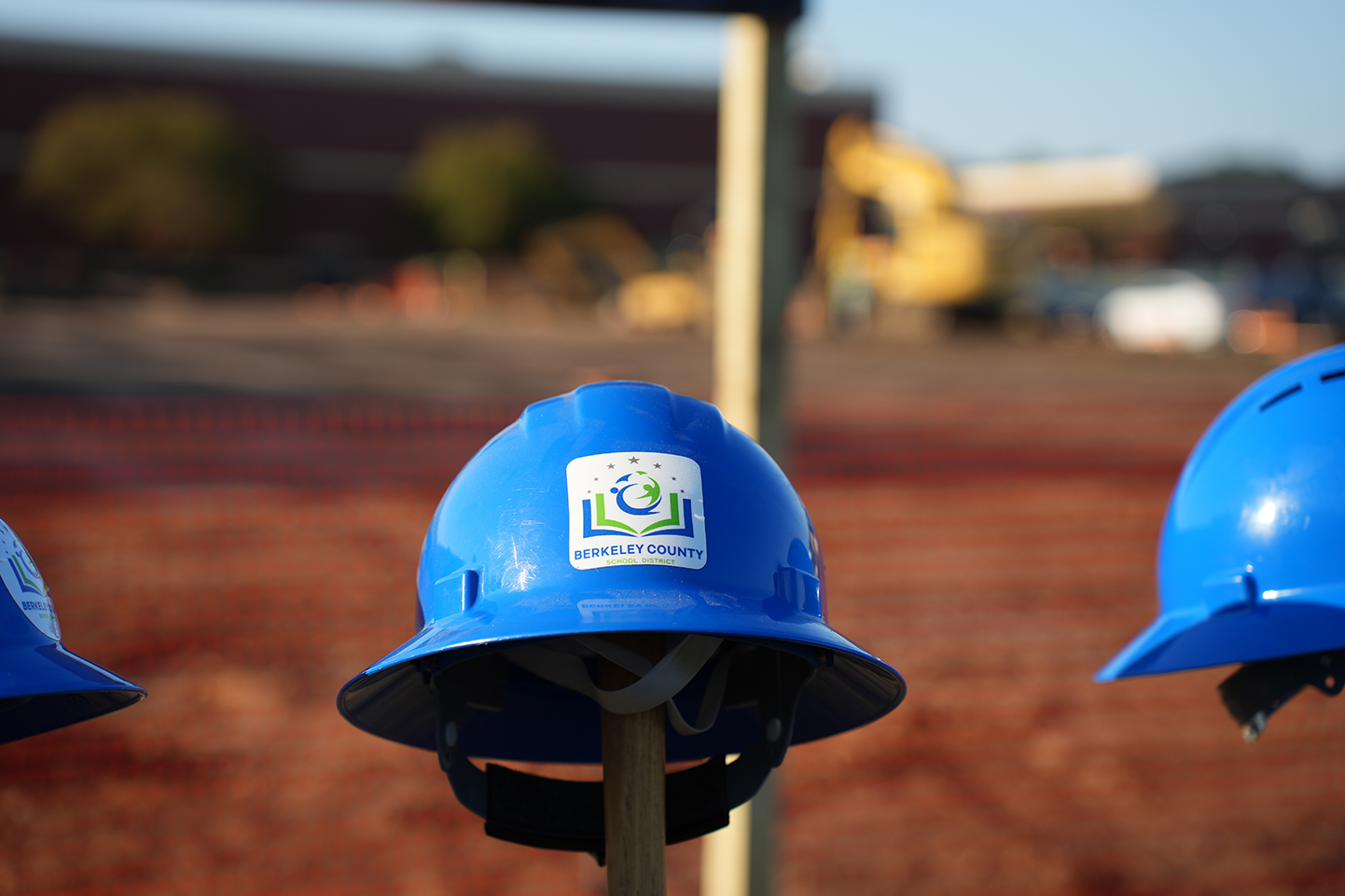 Three blue hard hats with reflective stickers displayed against a construction site backdrop with machinery and a red fence.