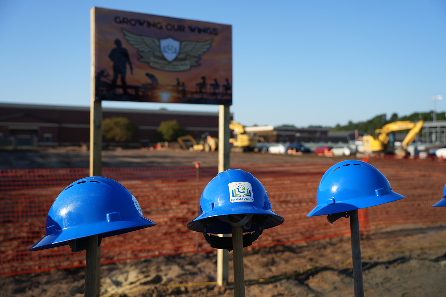 Four blue helmets stand on posts. Behind them, a sign reads "Growing Our Wings". Construction equipment is in the background.