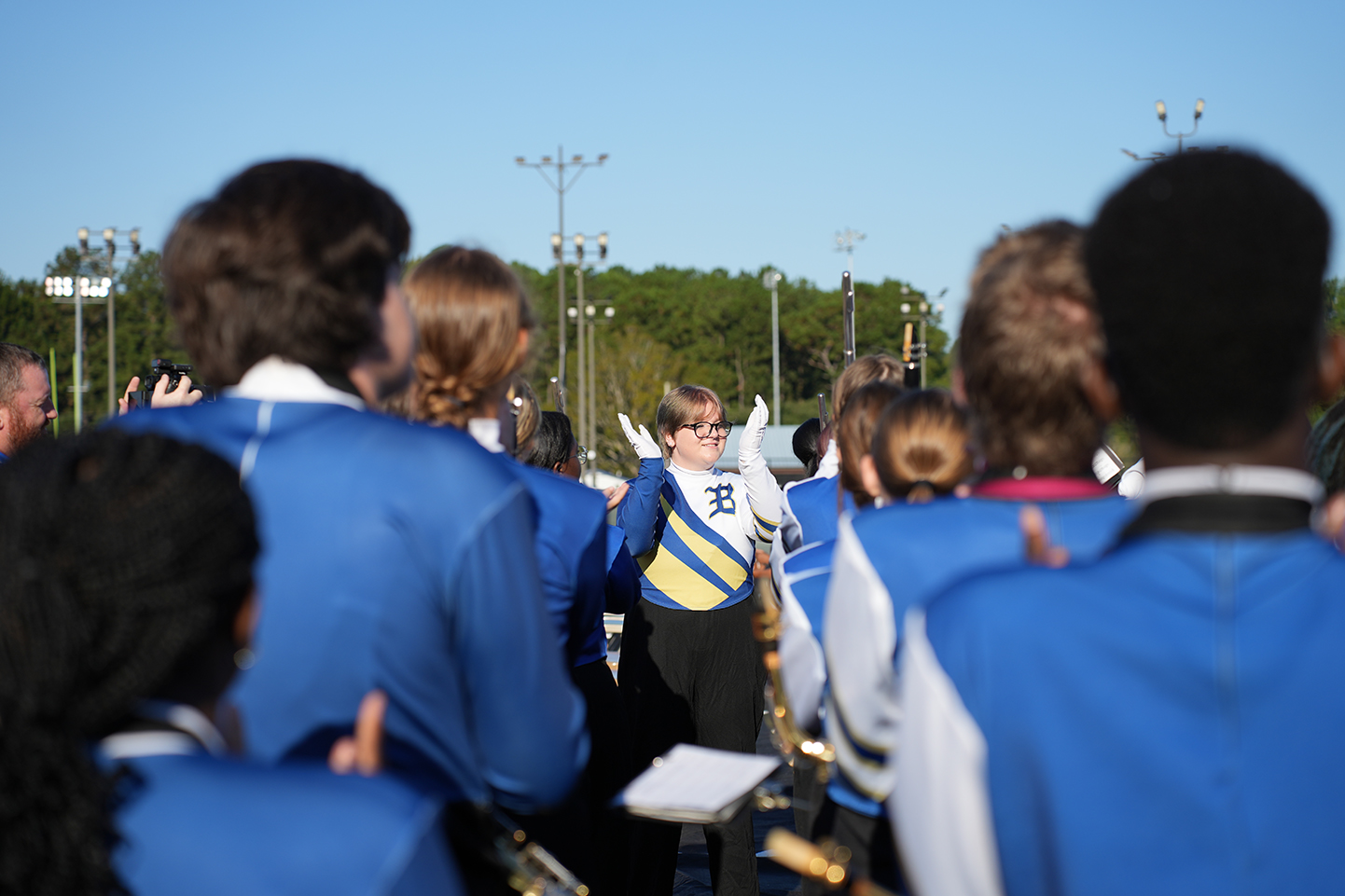 A group of people in blue and white uniforms, possibly a school marching band, standing outdoors with trees in the background.