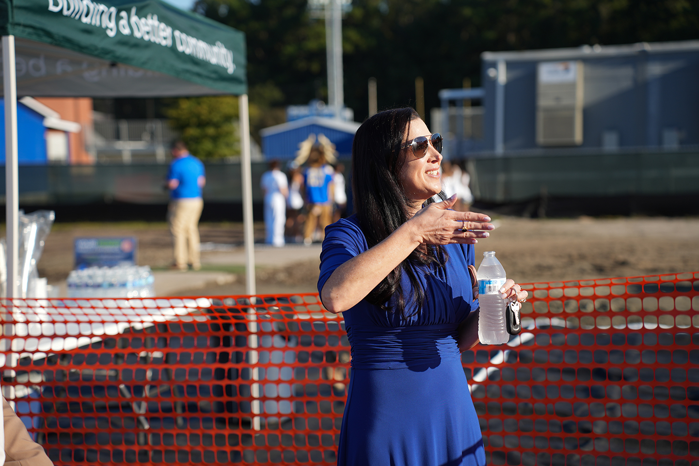 Person in blue dress stands at table, gesturing with hands while holding a water bottle.