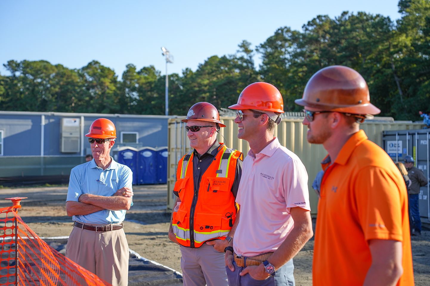 Five men in hard hats and short-sleeved shirts standing in a construction site. Trees in background.