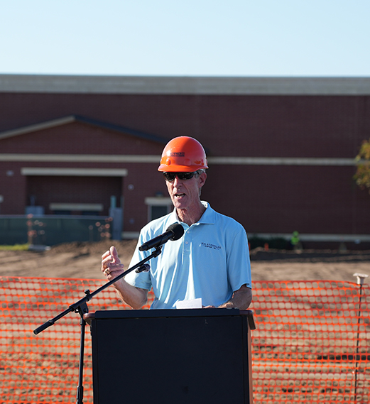 Person wearing orange hard hat speaks at podium with microphone in front of construction site.