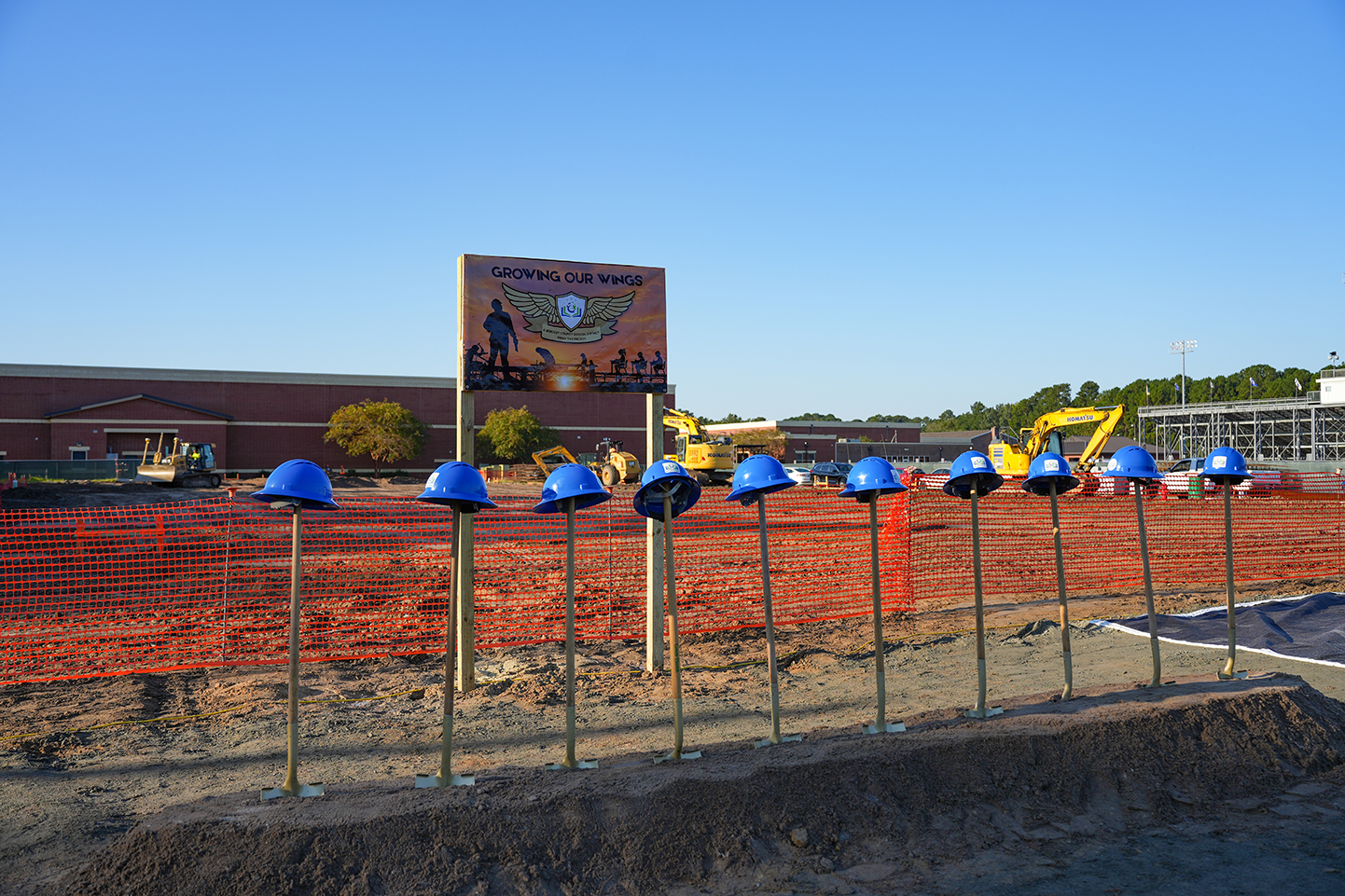 Row of blue helmets stands in dirt. Behind them, a sign, a construction site, and a building under construction.
