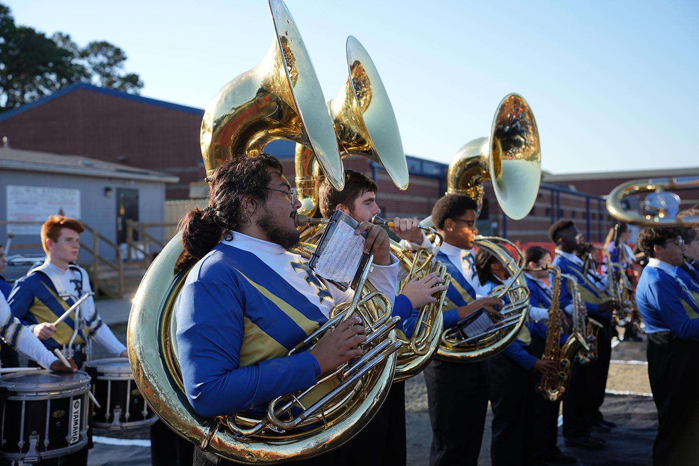 People in blue uniforms with large instruments play outdoors. One man holds a golden tuba. Behind, buildings are visible.