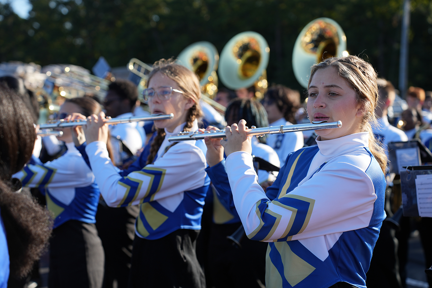 Three students in blue and white uniforms playing flutes, with others in a band behind them.