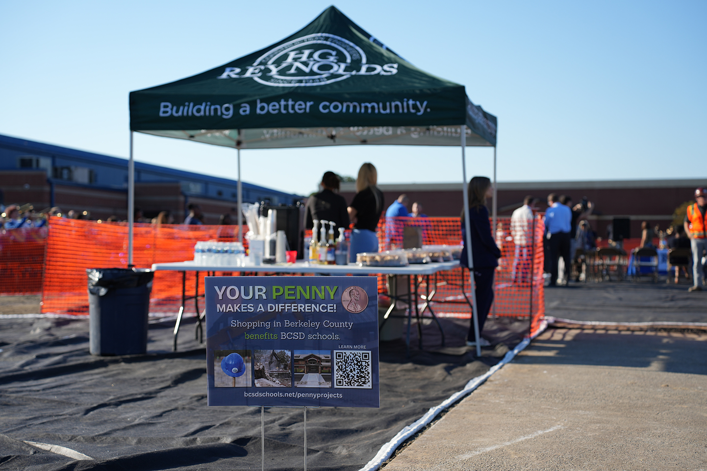 A tent with green and white sides displays a banner. A table holds a box, drinks, and snacks.