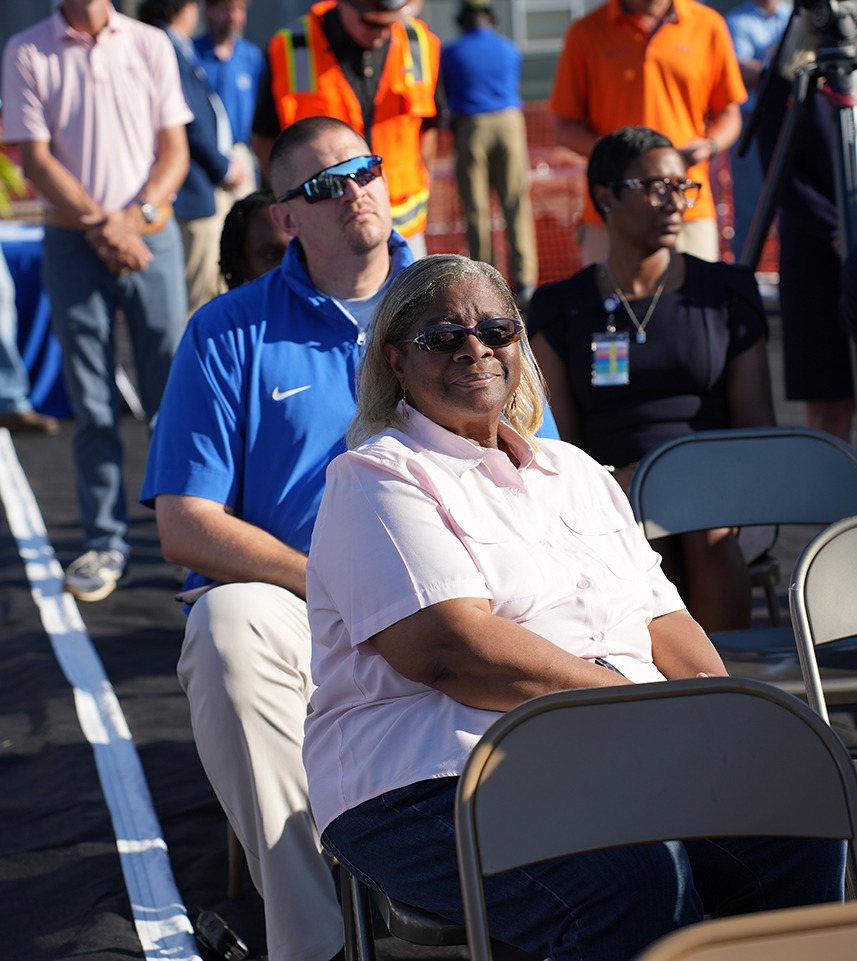Several people seated in chairs on a field. One woman in front wears glasses and a white blouse.
