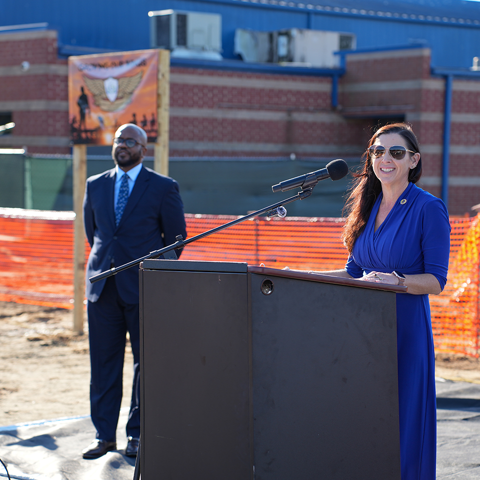 Two people at podium with microphone in front of construction site, one speaking, other standing.