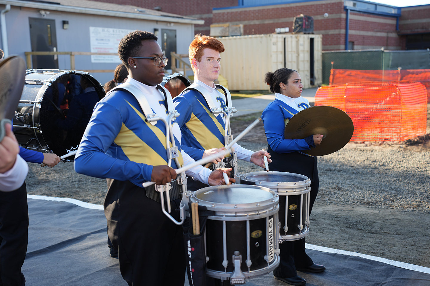 Three people in marching band uniforms playing drums and holding sticks with drumsticks, on a road.