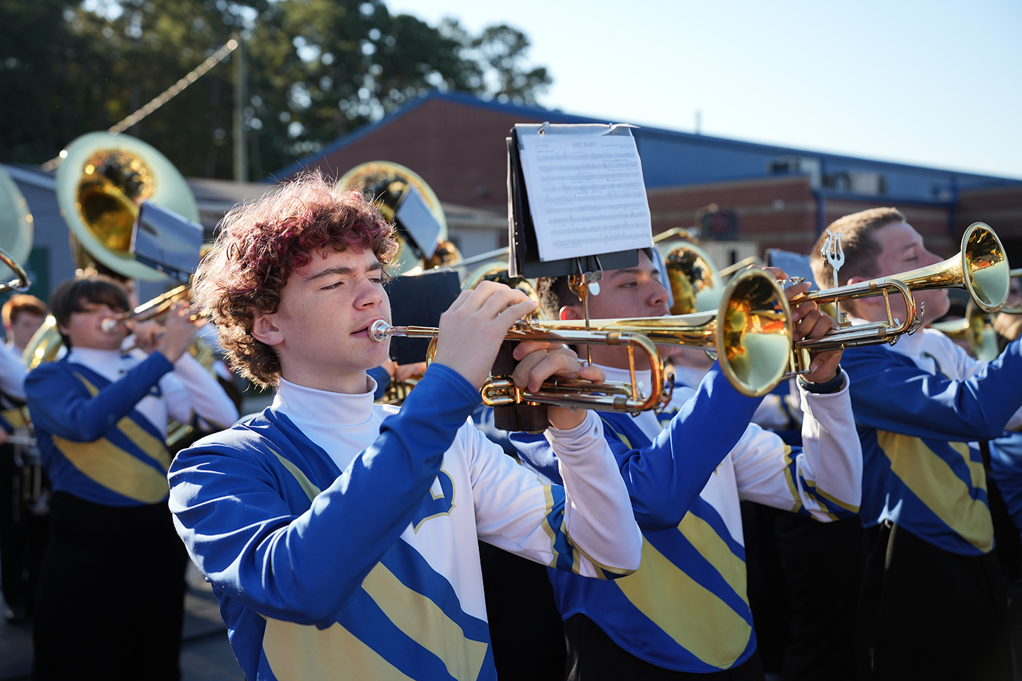 Several musicians in blue and yellow uniforms play brass instruments outdoors. A large building is visible in the background.