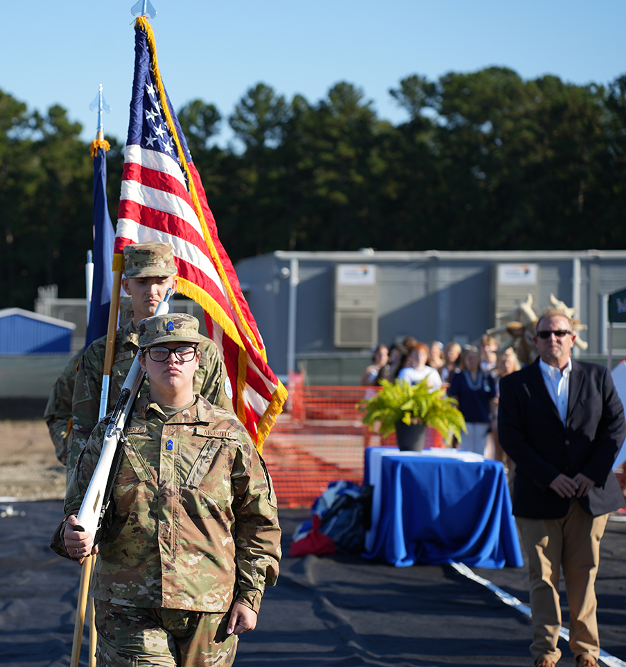 People standing outside, two in military uniforms, one holding flag, others watching, blue table with plant.