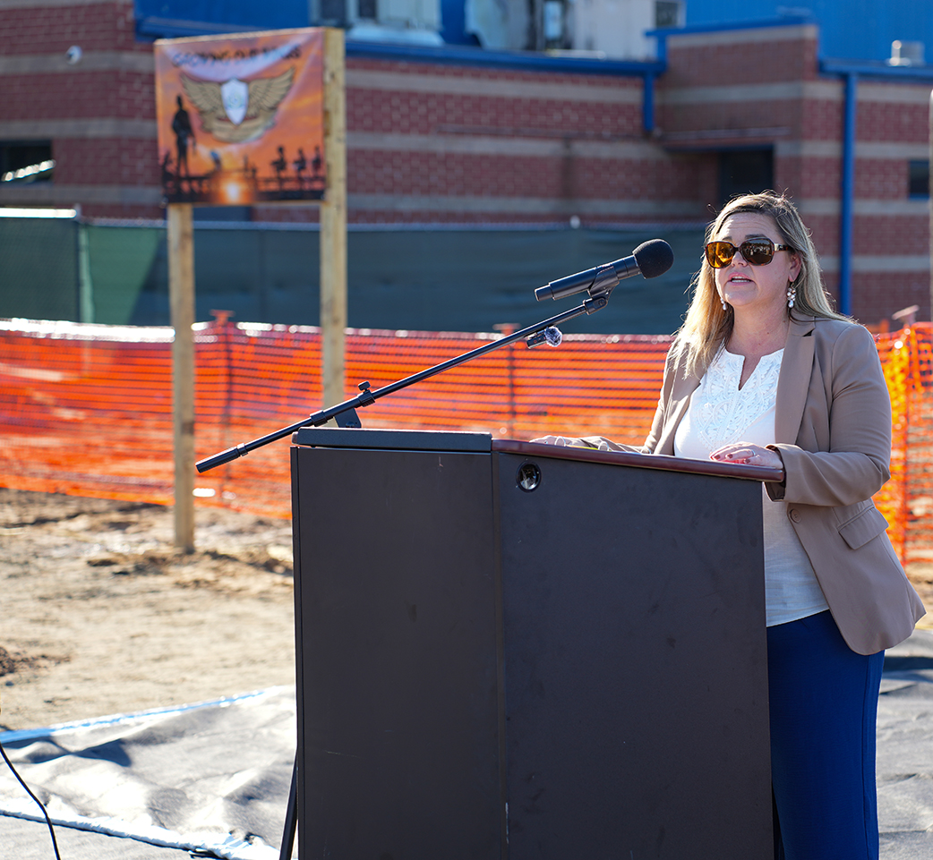 Woman at podium with microphone in front of construction site with orange fencing.