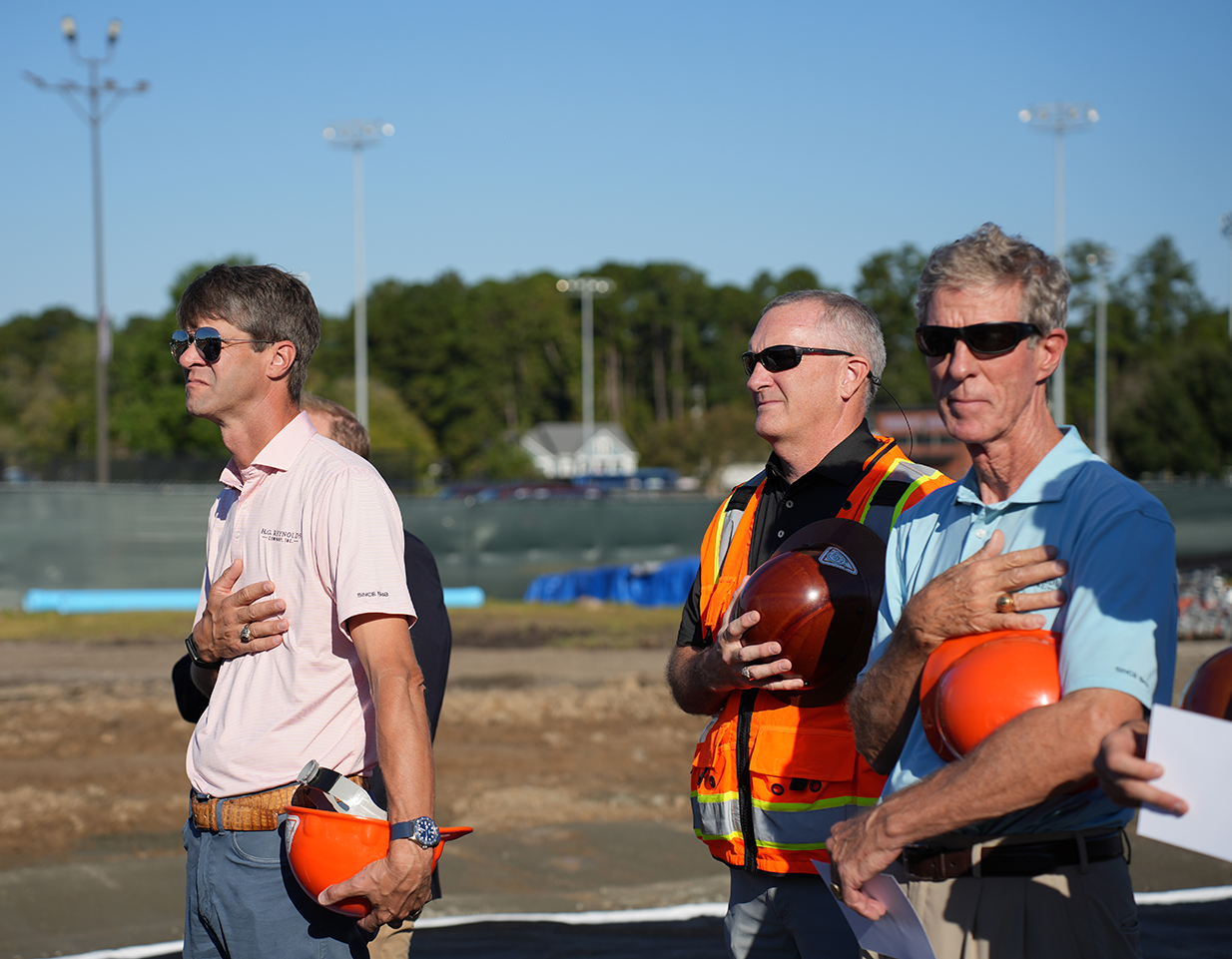 Four men stand on a construction site, one wearing a pink shirt and another an orange vest. Two men hold orange helmets, one holding a football. Trees and a house visible in the distance.