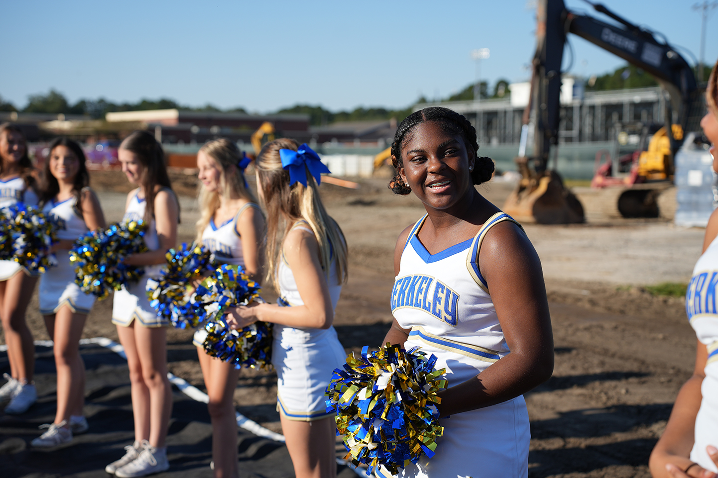 Six cheerleaders in matching uniforms stand in a row on a dirt field with a construction site in the background.