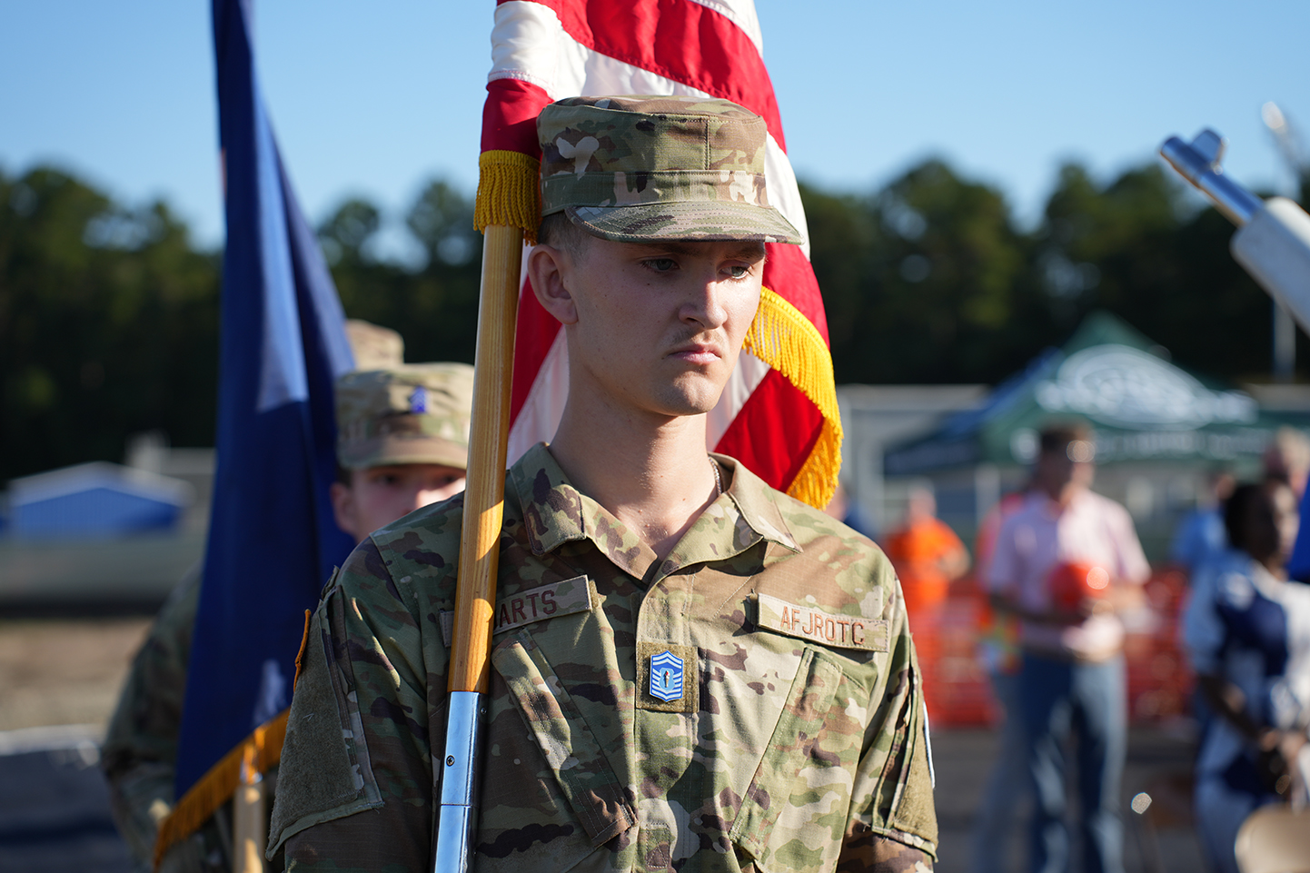Person in military uniform stands with flag in hand; others in uniform behind and to sides.