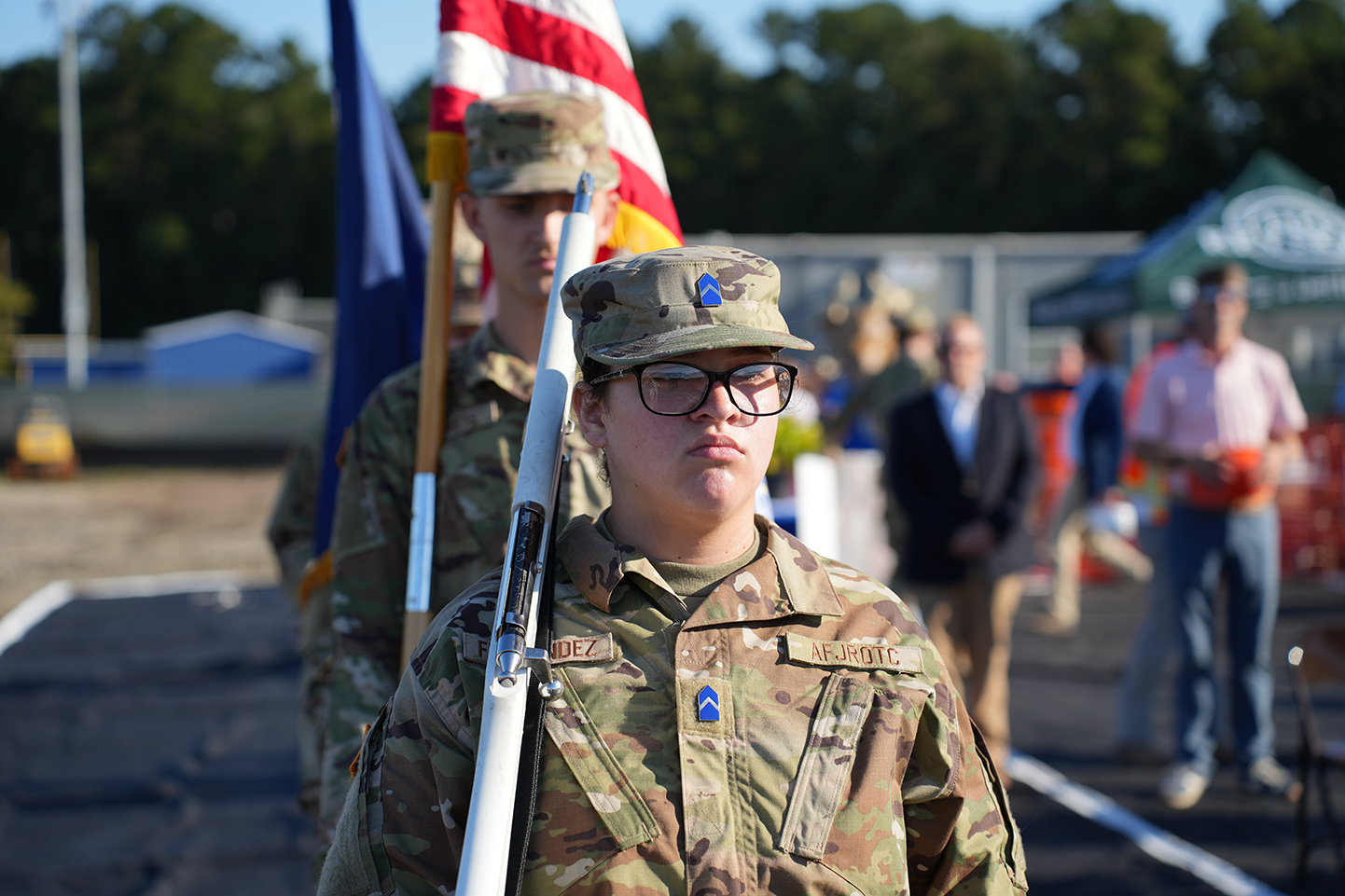 Soldier with glasses holds a rifle while standing with two other uniformed people. A flagpole with a flag behind them.