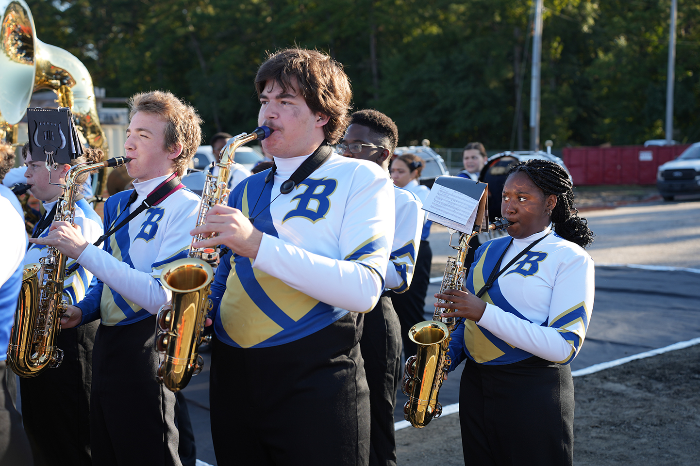 Six band members playing saxophones on a street, with trees and a building in the background.