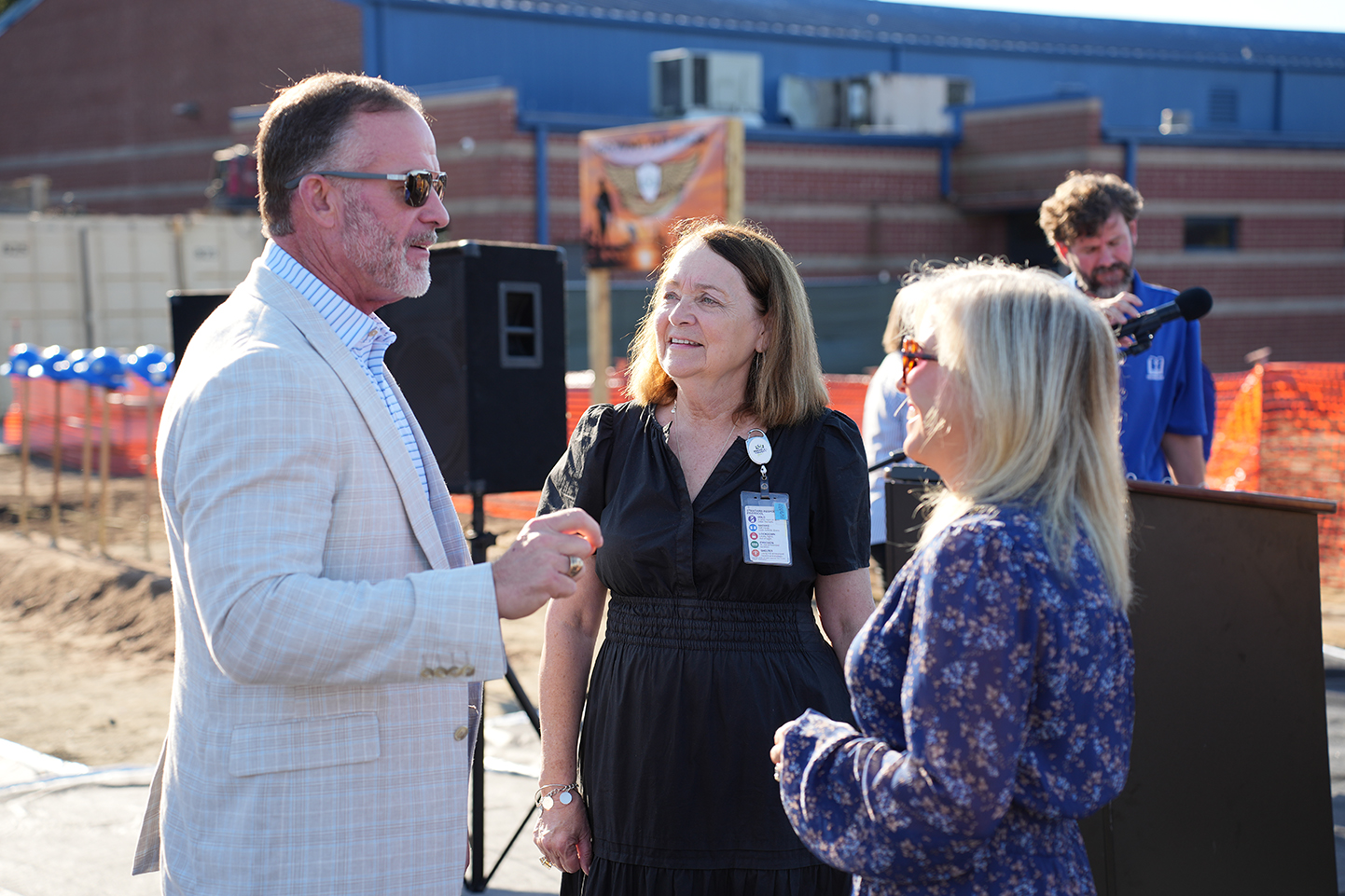 Four adults in formal attire stand in front of a podium with a microphone. A construction site is in the background.