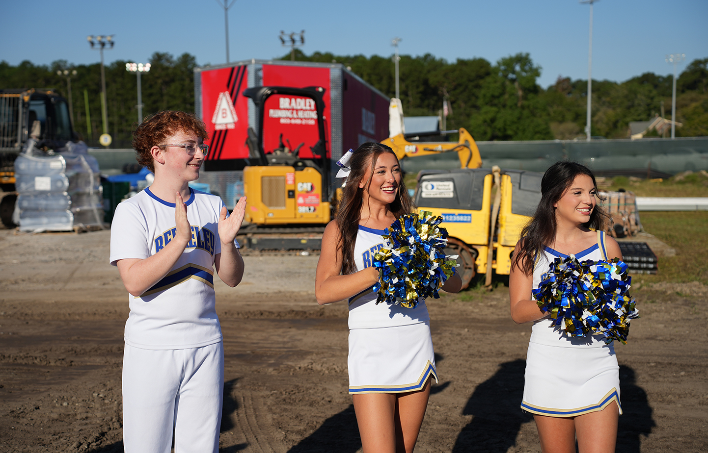 Three young women in cheerleading uniforms standing on a dirt field with construction equipment in background.