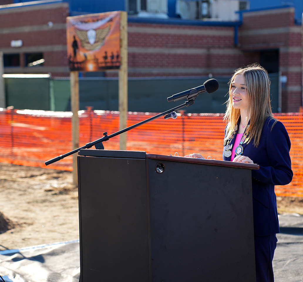 Person with microphone at podium, standing beside a barrier, with a building and sign in the background.