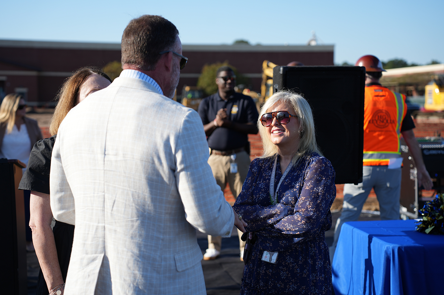 Adults standing outdoors with blue table. Man and woman talking. Man in white suit and sunglasses. Woman in floral shirt and sunglasses.