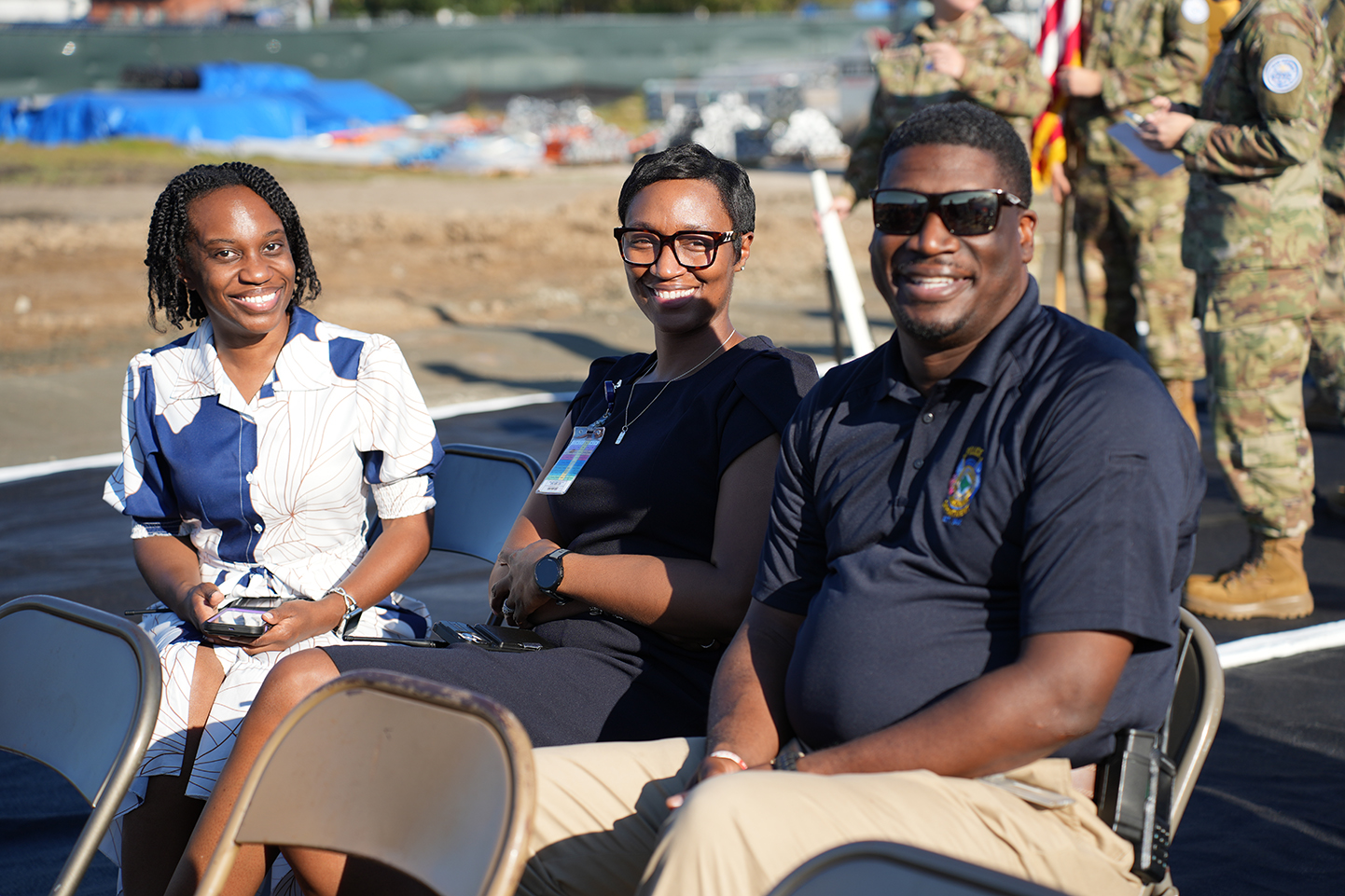 Three adults in chairs outdoors. One woman holds a device. Two men in uniform stand behind them.