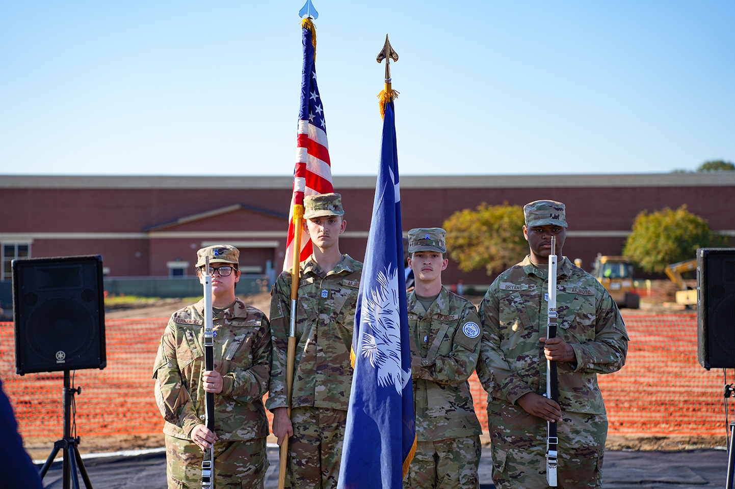 Four service members in uniform stand in a row, holding flags and rifles. Behind them, a building, trees, and dirt area are visible.