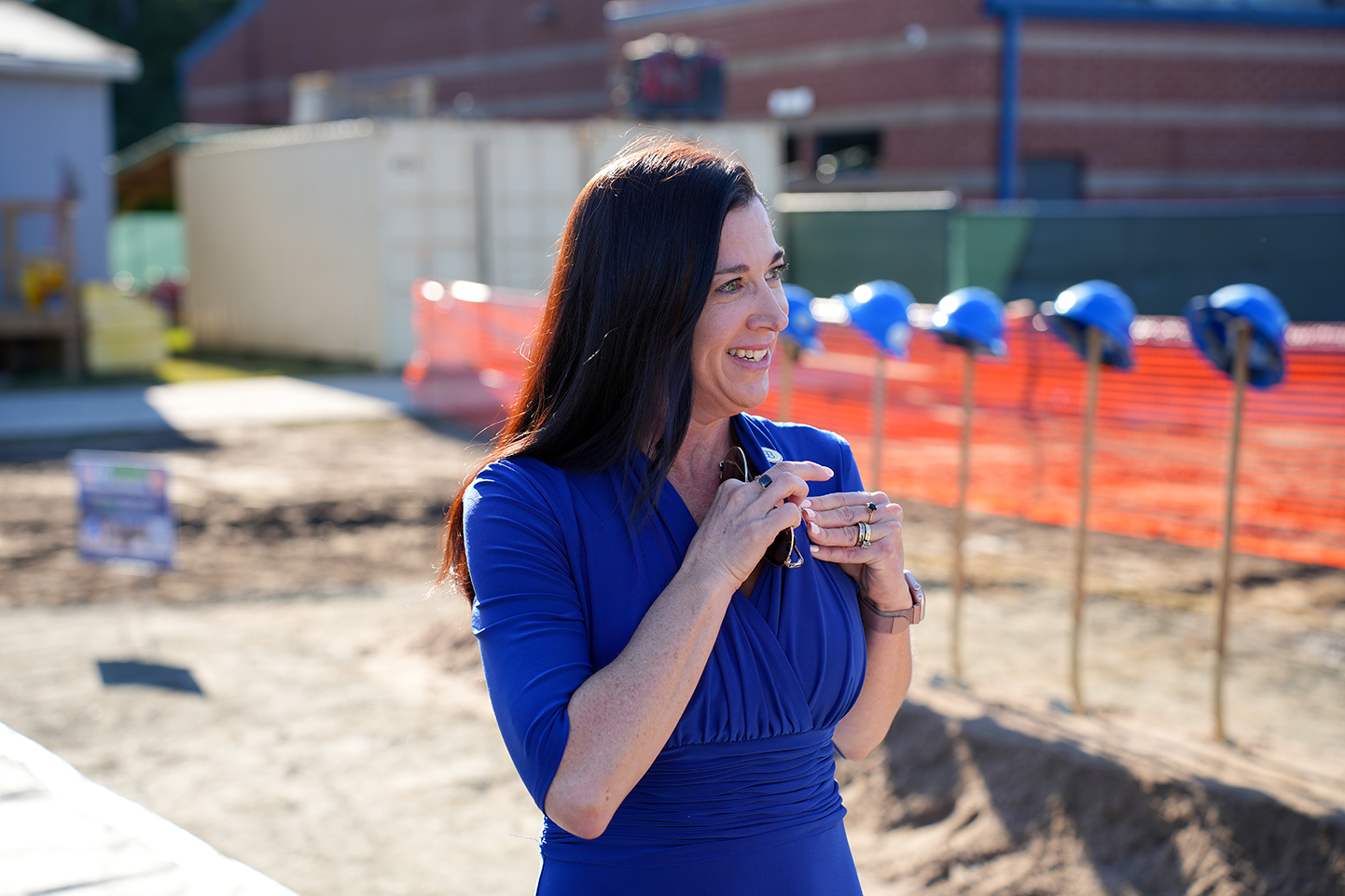 Woman standing in front of a building and smiling, holding a cup, with hard hats in a row behind her.