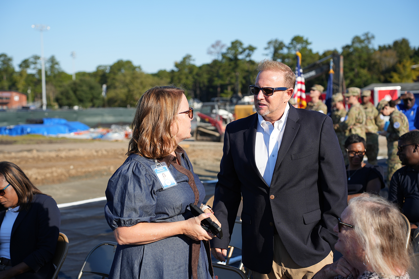 People converse at an event. A woman holds a camera. A man in glasses stands with his arms folded.