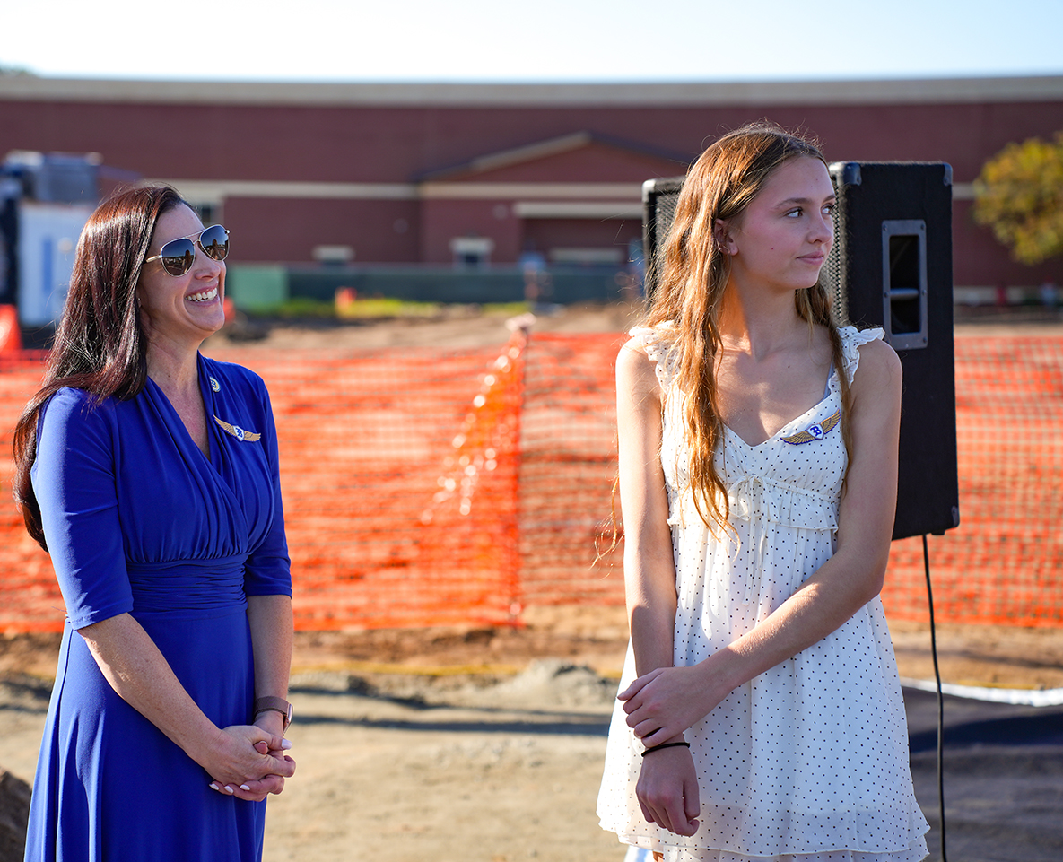 Two women, one in a blue dress and the other in a white dress, stand by a fence. Behind them is a building and a speaker.