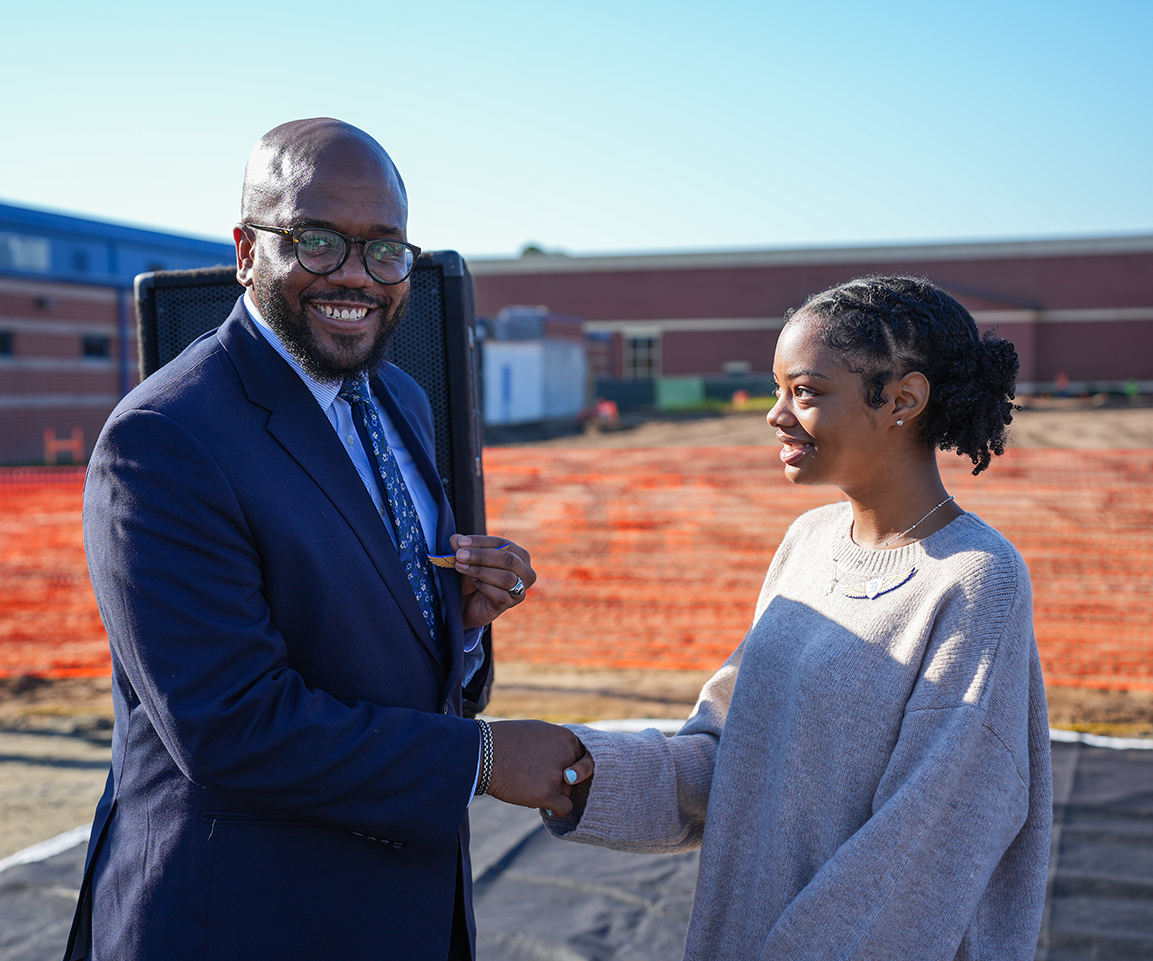 A man and woman shake hands in front of a school building. Behind them, construction work is ongoing.