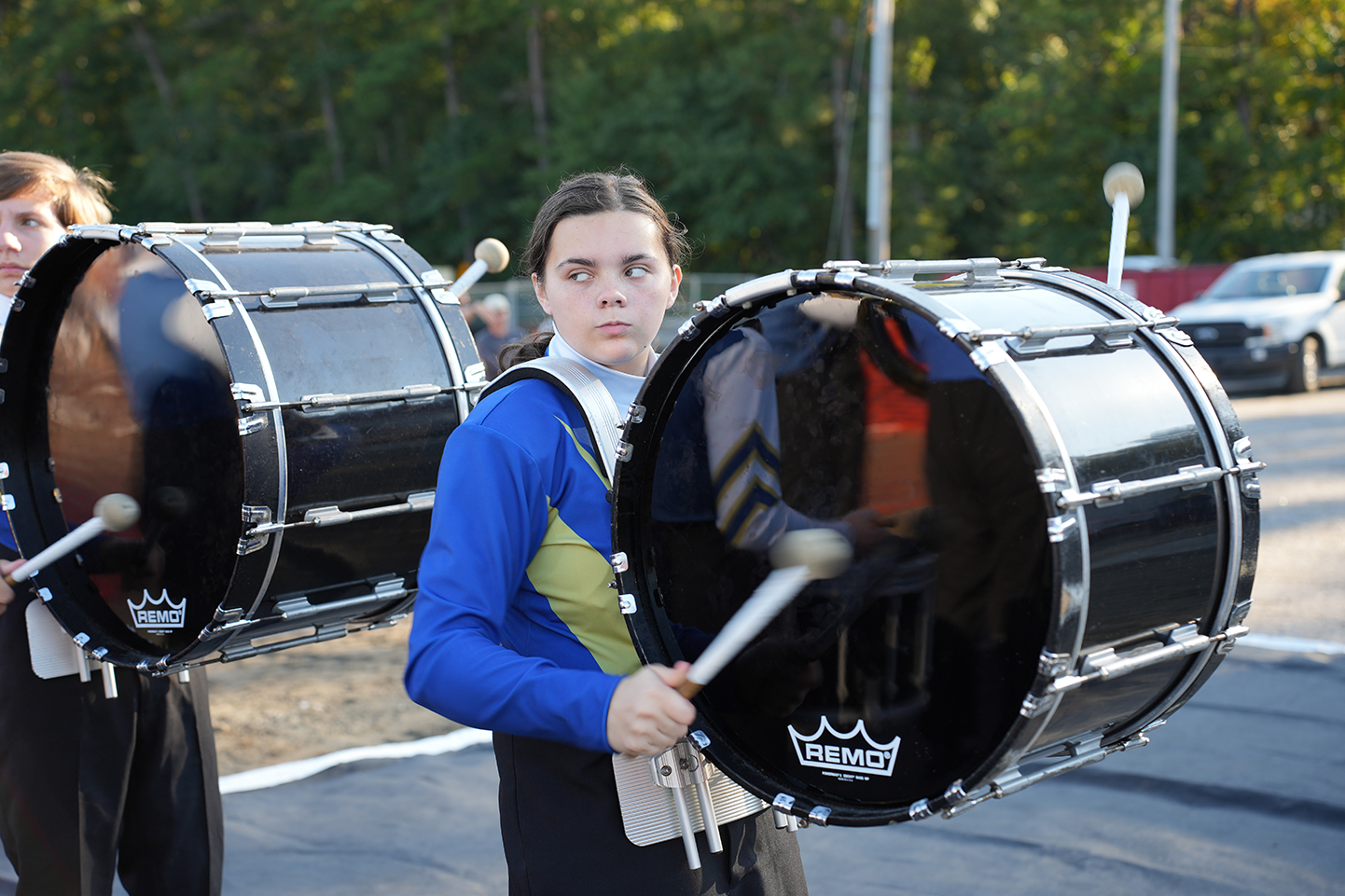 Two students with drumsticks hold large black drums. They are wearing blue uniforms. Trees and a car are in the background.