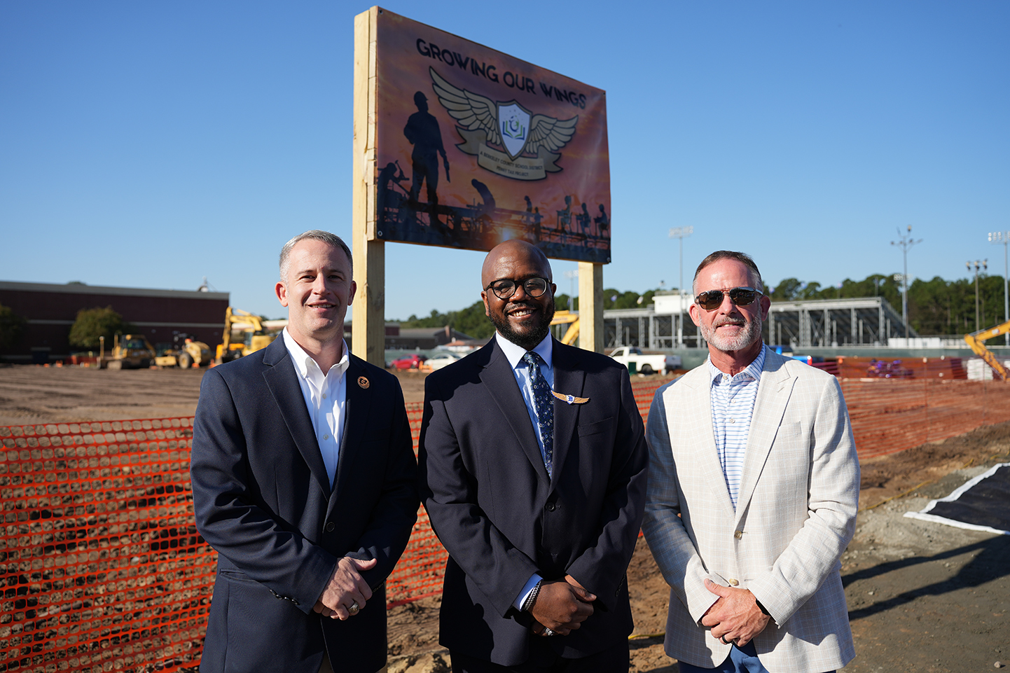 Three men in business attire stand in front of a construction site, near a sign with a logo.