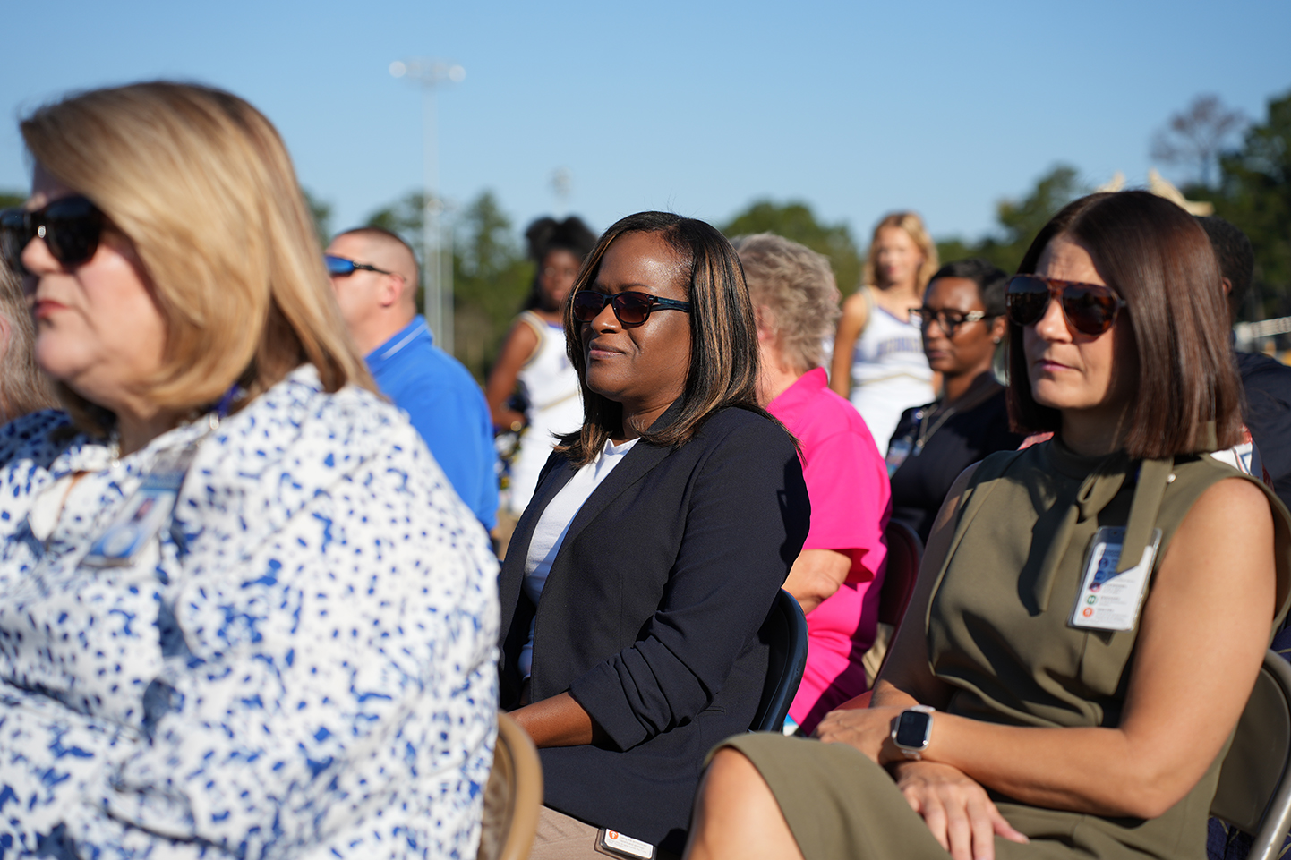 Several adults wearing sunglasses and seated in chairs facing the same direction, with bleachers in background.