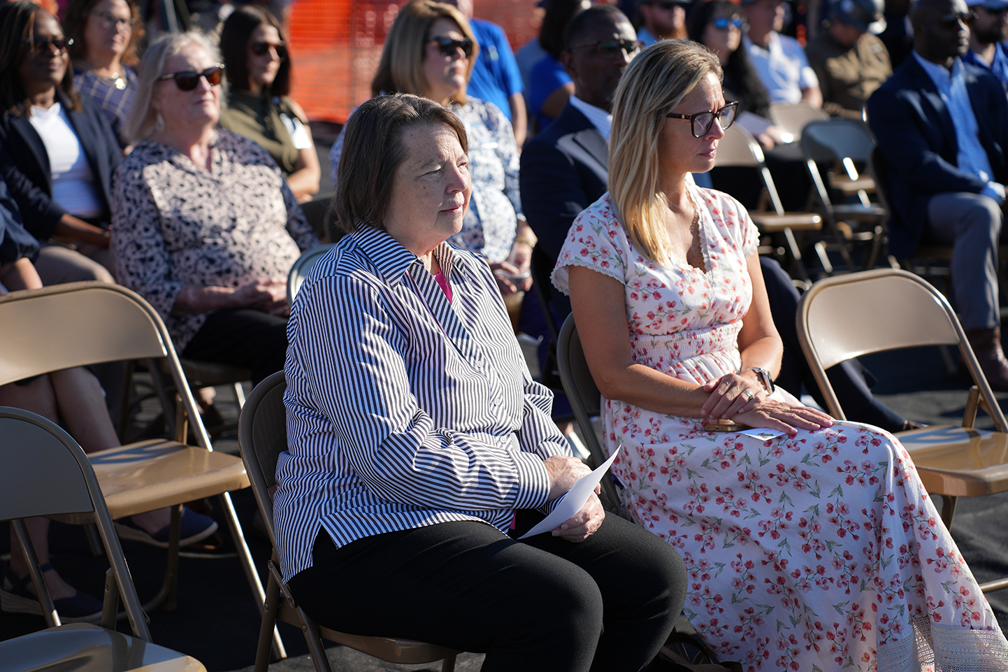 People in rows of chairs facing forward. One woman holds a paper, another wears glasses, and two women wear dresses.