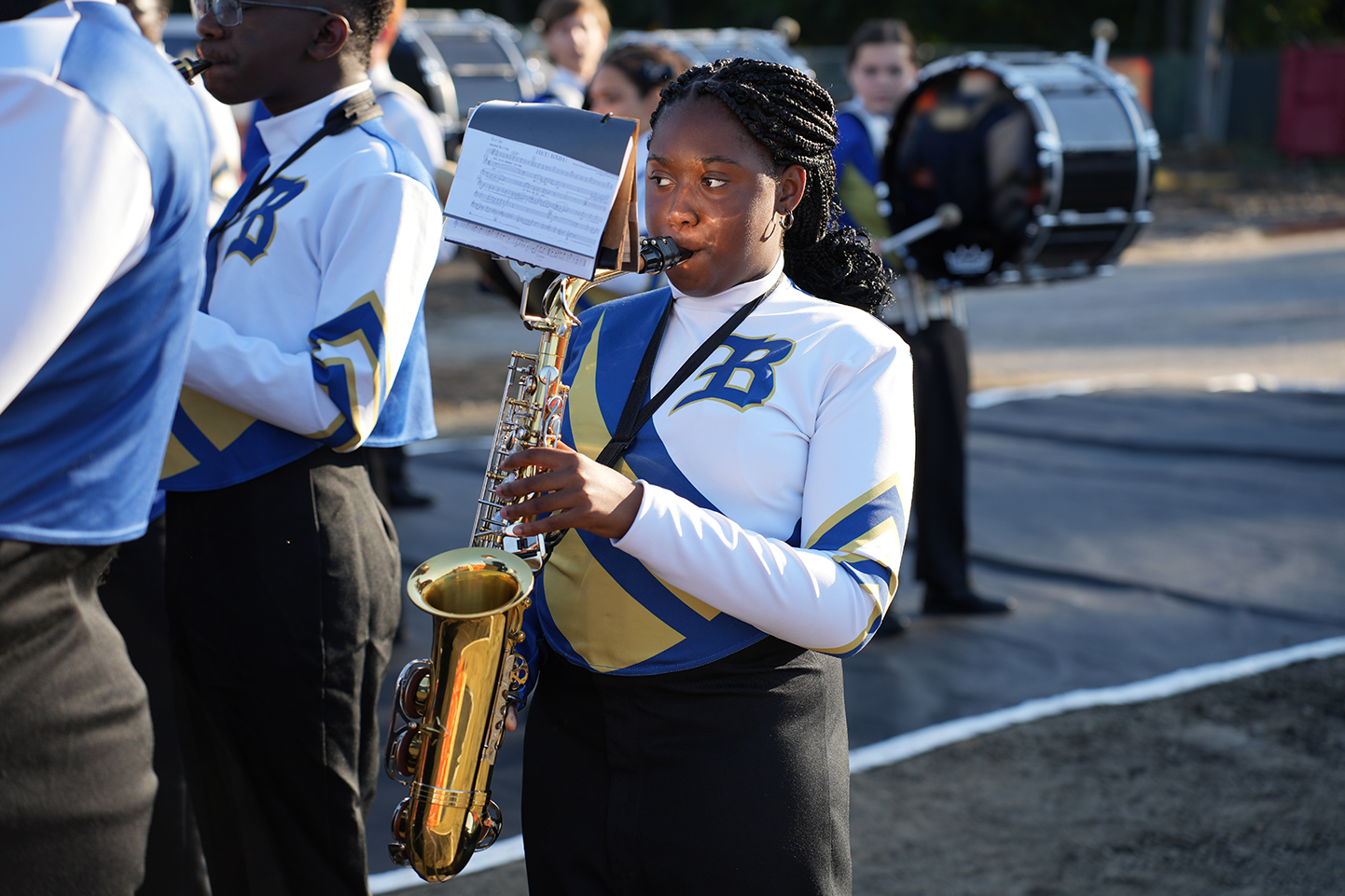 A student stands in a group, holding a saxophone, with other students and instruments in the background.